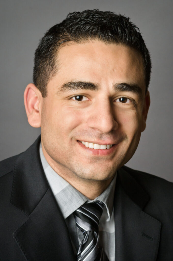 Headshot of a man in his 20s who has heritage from Oman with light skin tone and black short hair. He is wearing a black suit, white and grey checkered button up shirt with a black and grey tie. He is smiling at the camera.