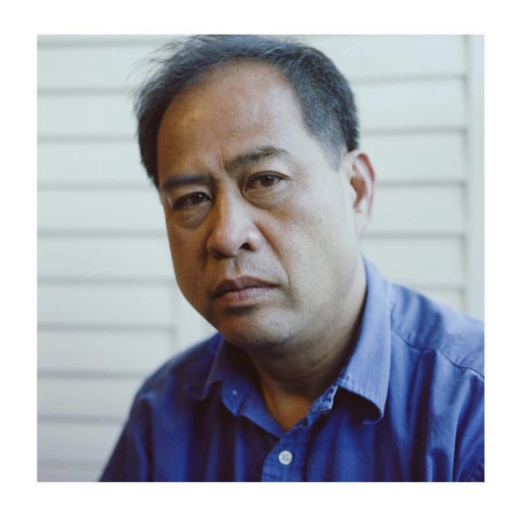 Photograph of a man in his 40s with medium skin tone and black balding hair. He is wearing a blue button up shirt and there is a white slatted wall behind him.