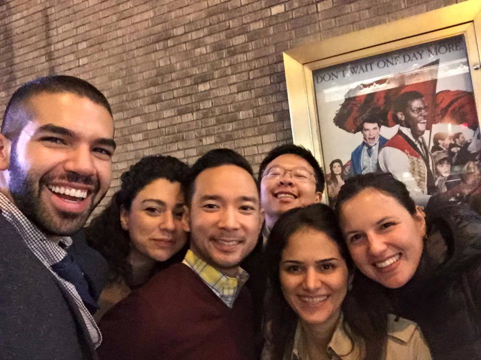 Six people of varying heritages, all in their 20s, stand in front of a poster for "Hamilton" the musical on Broadway. 