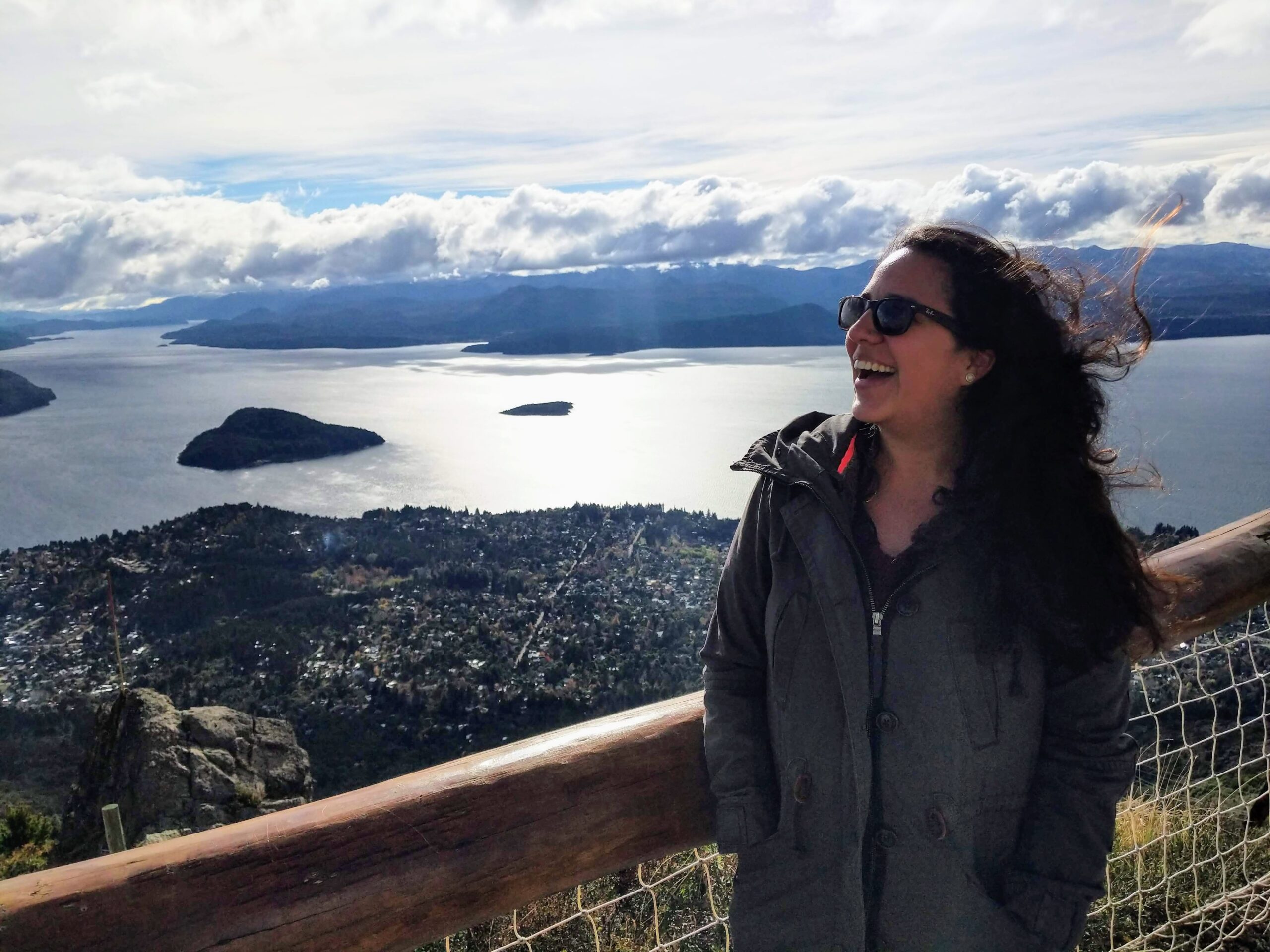 Photograph of a woman standing at a handrail overlooking a mountainous, water-filled landscape. She is wearing a jacket while the wind blows her hair and she is wearing sunglasses and laughing.