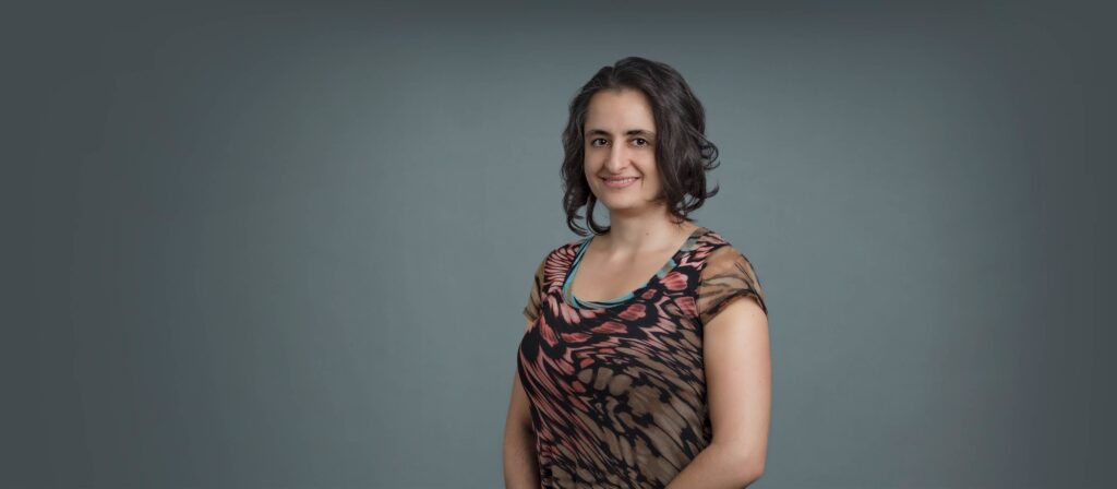 Headshot of a woman in her 20s with heritage from Ukraine, she has light skin tone and wavy dark brown long bob cut. She is wearing a multi-colored short sleeve shirt and smiling at the camera.