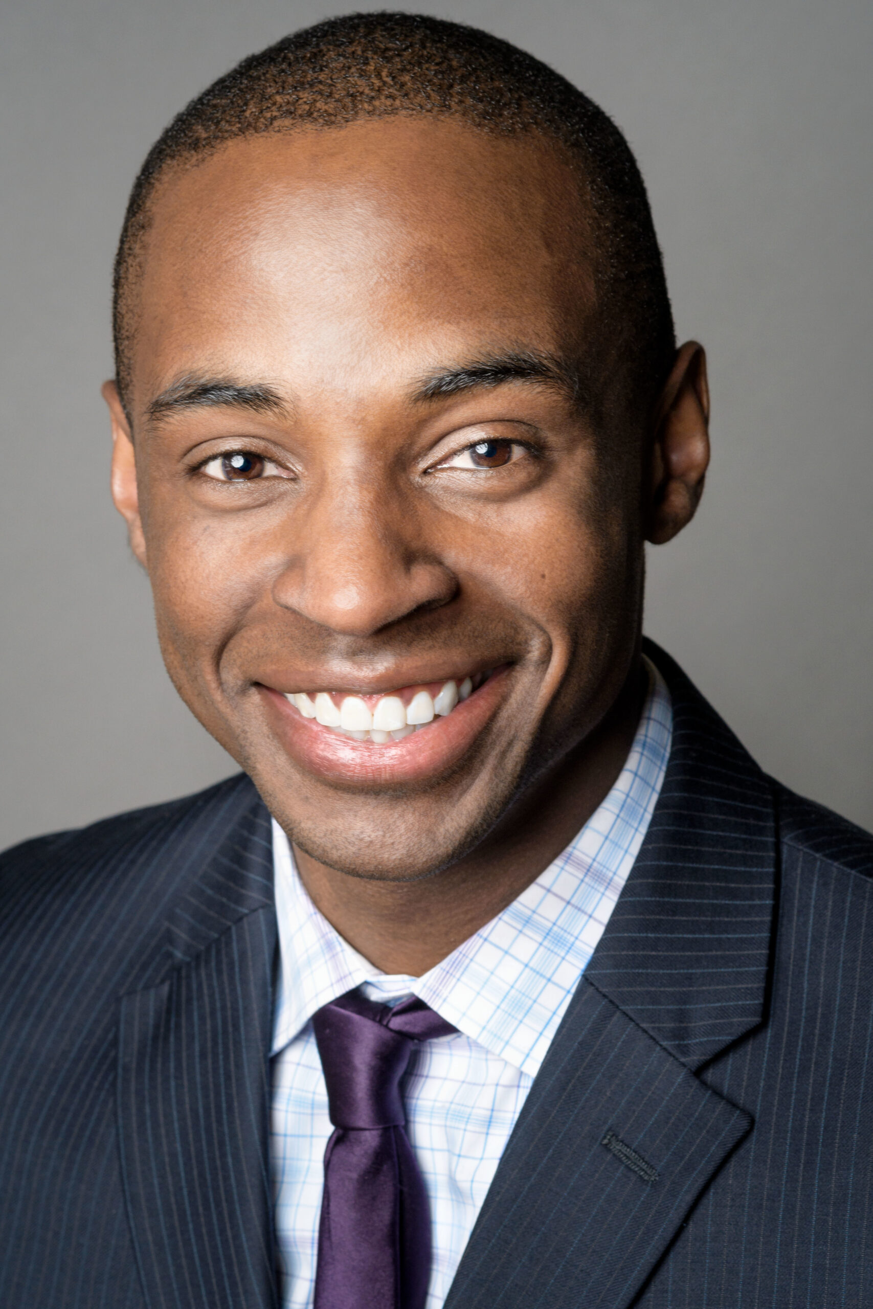 Headshot of a man in his 20s with heritage from Nigeria he has dark-medium skin tone and buzzed black hair. He is wearing a navy pinstripe suit, white and blue gingham button up and purple tie. He is smiling at the camera.