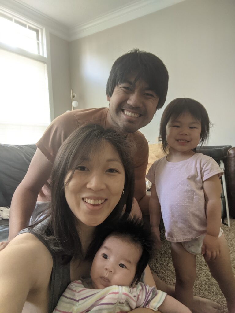 Selfie of a family, mom, dad and two young children. They are cheesing for the camera in what appears to be a living room. 