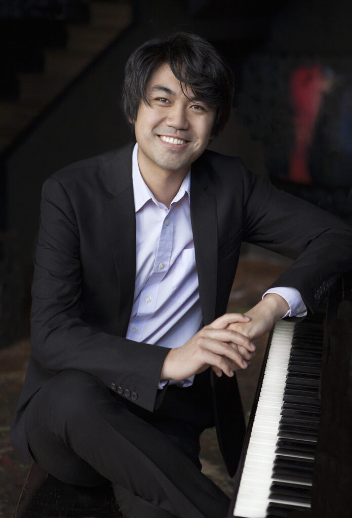 Headshot of a man in his 30s who has heritage from Taiwan with light skin tone and shaggy black hair. He is wearing a black suit and white button up. He is resting his left arm on a piano and smiling at the camera. 