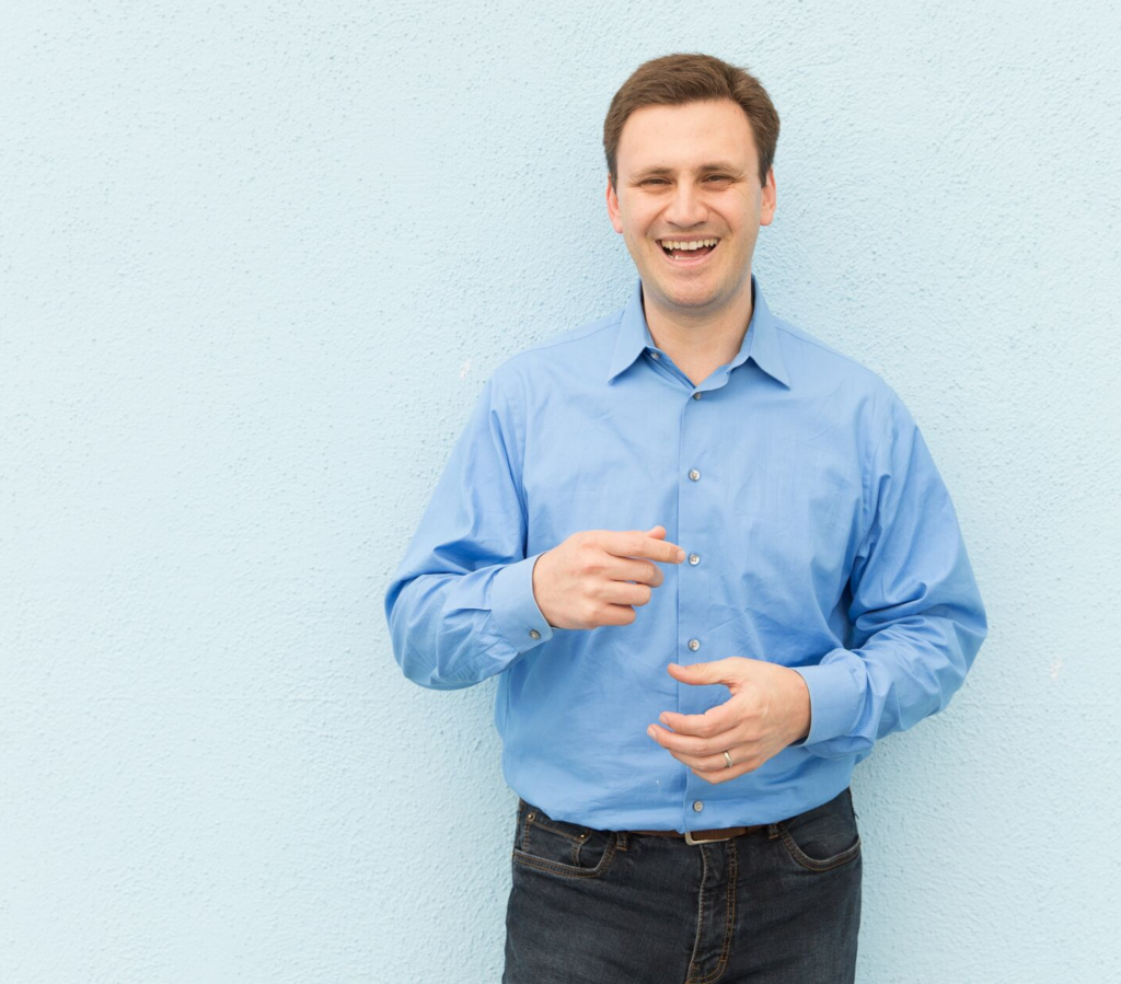 Half body photograph of a man in his 30s who has heritage from Czech Republic with light skin tone and light brown crew cut hair. He is wearing a blue button up shirt and dark jeans. He is standing against a light blue wall, gesturing with his hands and smiling big at the camera. 