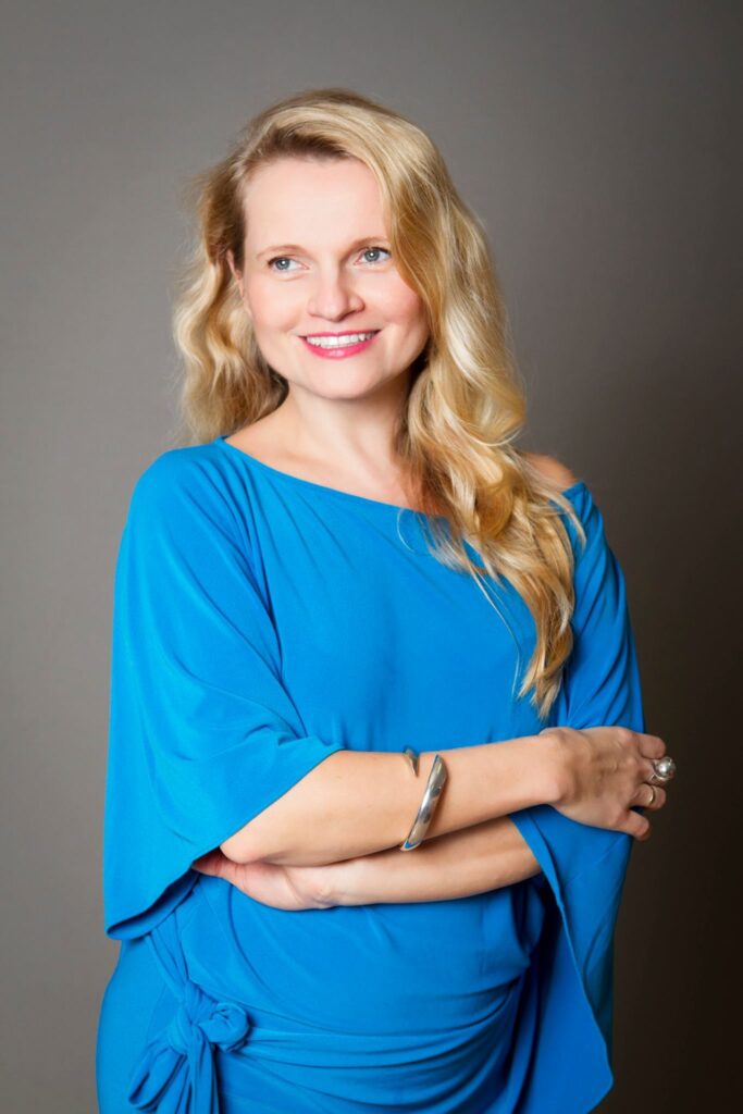 Headshot of a woman in her 30s with heritage from Poland she has light skin tone and wavy, long blonde hair. She is wearing a bright blue boat neck dress, bracelet and a few rings. She is standing with her arms crossed, looking slightly off camera left and smiling.