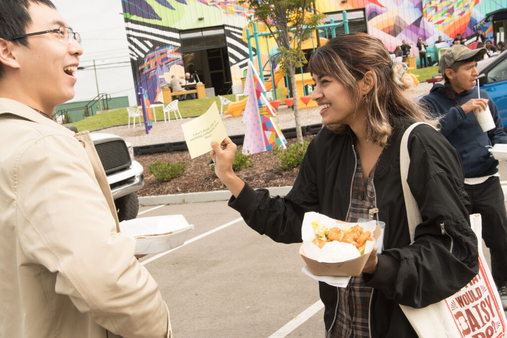 Photograph of a couple people in their 20s, standing in a parking lot, talking and holding food. 