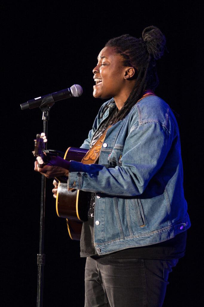 Photograph of a woman who has medium-dark skin tone and shoulder length braided hair. She is wearing a jean jacket, black t-shirt and black jeans. She is holding a guitar and singing into a microphone. 