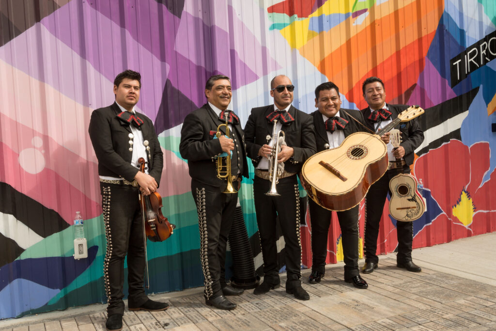 Photograph of a mariachi band, in full regalia, standing in front of a multi-colored mural wall. 