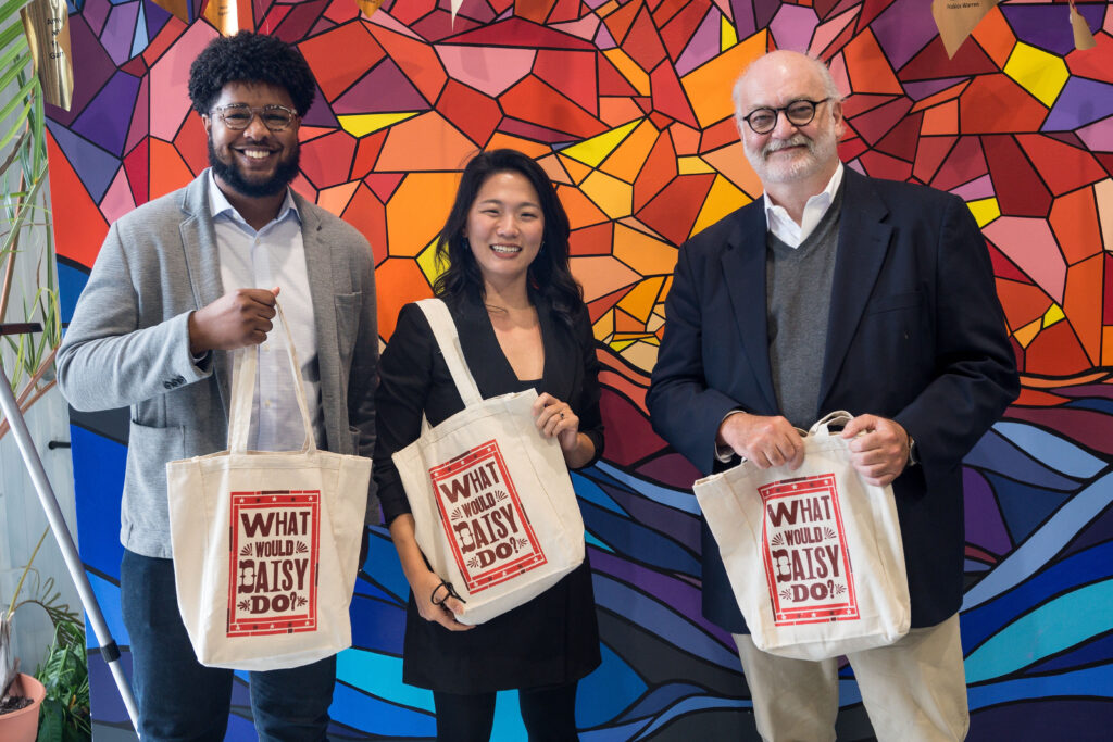 Photograph of three people posing with tote bags that say "What would Daisy do?", they are standing in front of a multi-colored wall. 