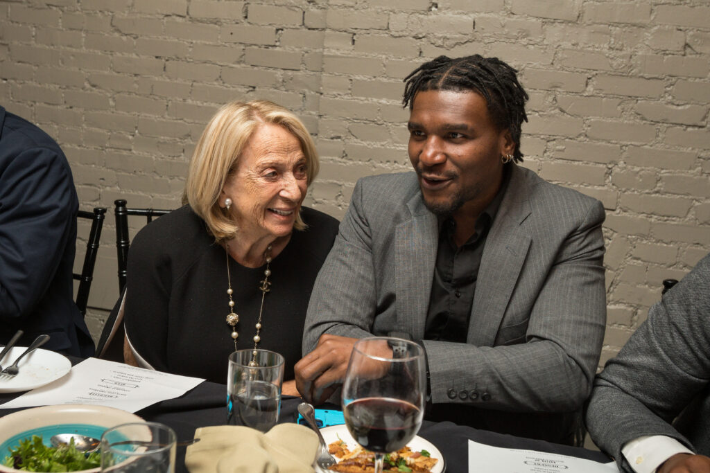 Photograph of two people at a dining table, the woman on the left is in her 80s, the man on the right is in his 20s. They are laughing. 