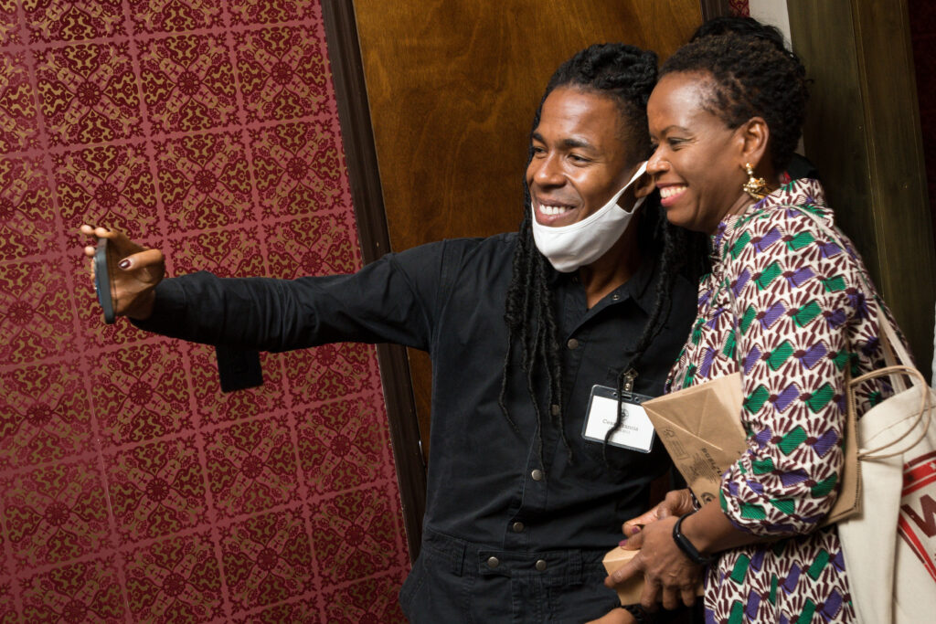 Photograph of two people taking a selfie. Behind them the walls are an intricate red pattern and wood. 