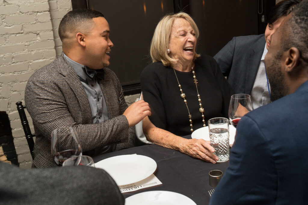 Photograph of four people sitting at a dining table. Three of the men are in their 20s, the woman at the center of the photo is in her 80s, they are all laughing. 