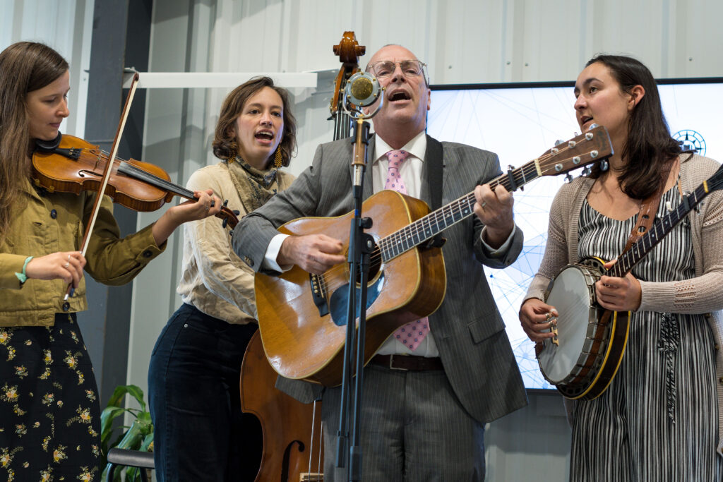 Photograph of a man playing guitar and singing at a microphone. Three women are surrounding him, the one on the left is playing a violin, the one in the back is playing an upright bass and on the right has a banjo. 