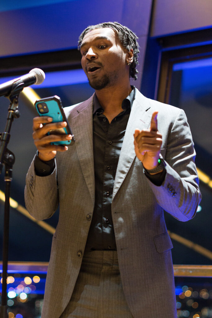 Photograph of a man in his 20s with medium-dark skin tone and black hair. He is wearing a grey suit and black button up shirt. He is holding a cell phone, speaking into a microphone and gesturing with his other hand. 