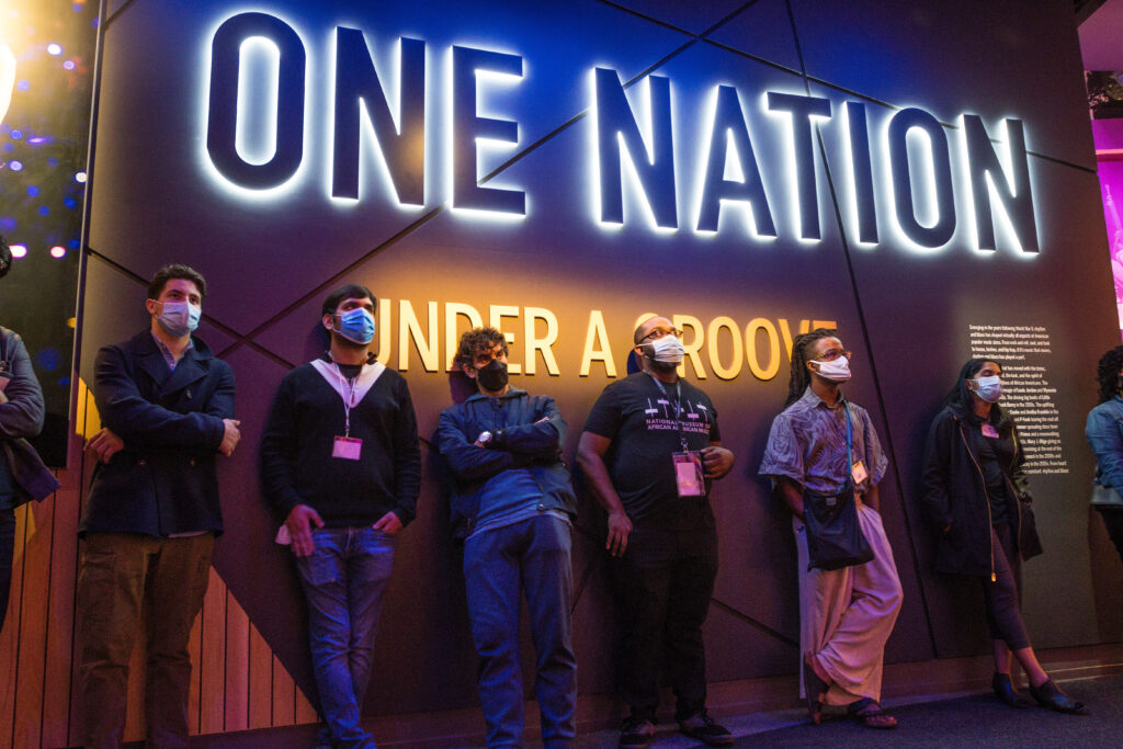 Photograph of people standing against a wall, they are wearing face masks. The wall they are leaning against says "One Nation under a Groove".