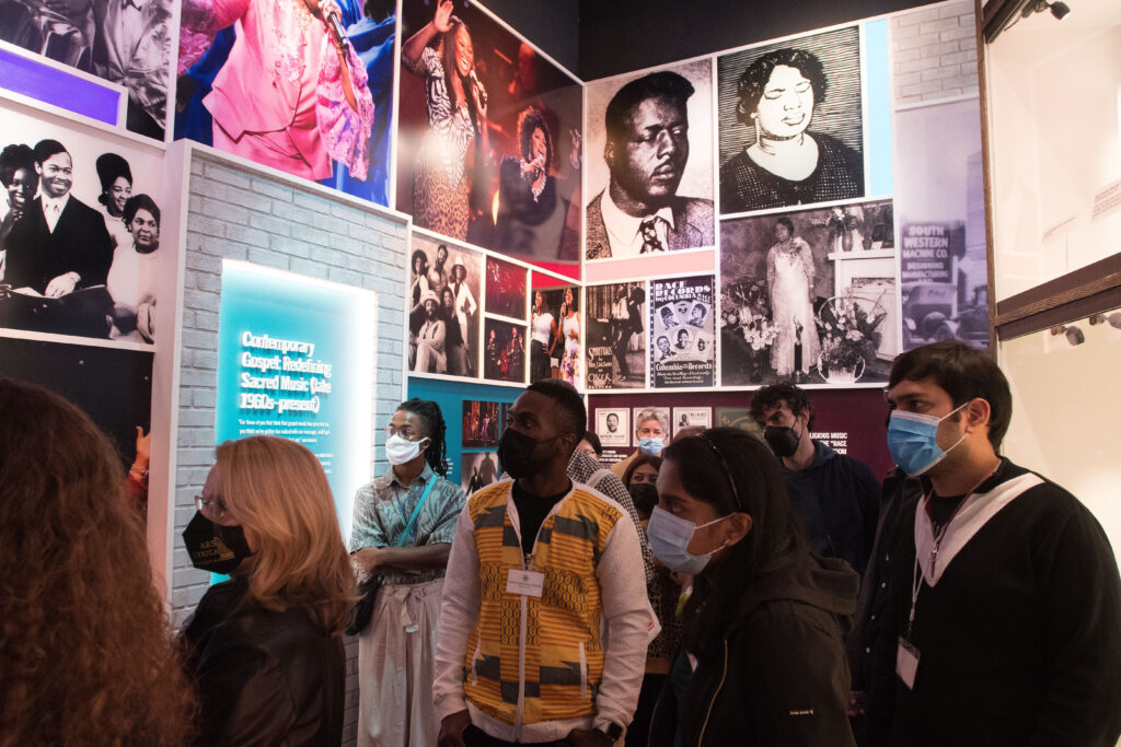 Photograph of a group of people in their 20s walking through a museum, they are wearing face masks. Old photographs and explanations can be seen on the walls behind them. 