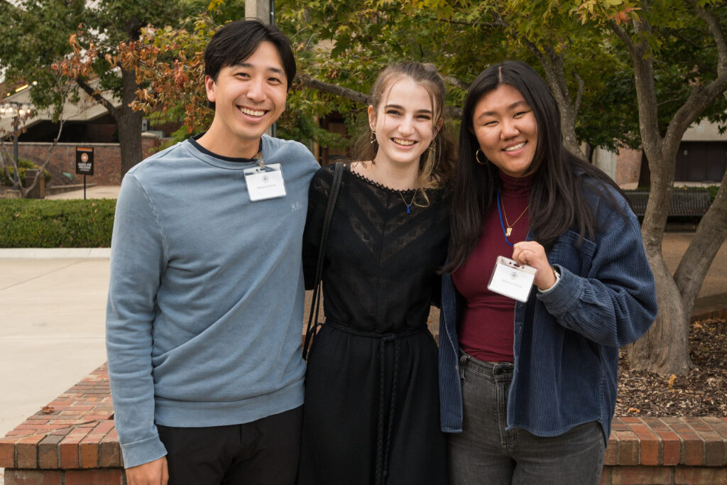 Photograph of three people posing for the camera, some trees and building can be seen behind them. 