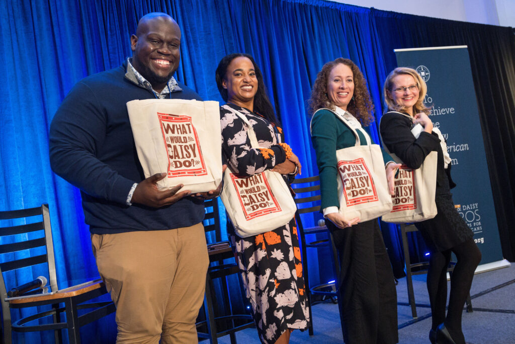 Photograph of four people posing with tote bags that say "What would Daisy do?". One man on the left and three women, all in their 20s of varying heritages. 