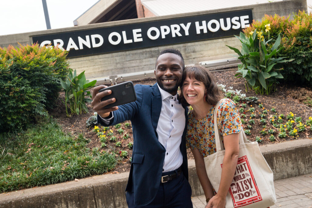 Photograph of two people taking a selfie in front of the "Grand Ole Opry House" sign. The man on the left is in his 20s, the woman on the right is in her 30s.