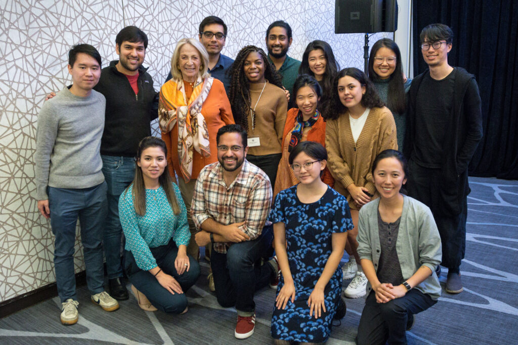 Photograph of a group of fifteen people posing for the camera. Most of the people are in their 20s with a variety of heritages. One woman is in her 80s. Behind them is a white wall with grey lined pattern across it and the carpet is blue.