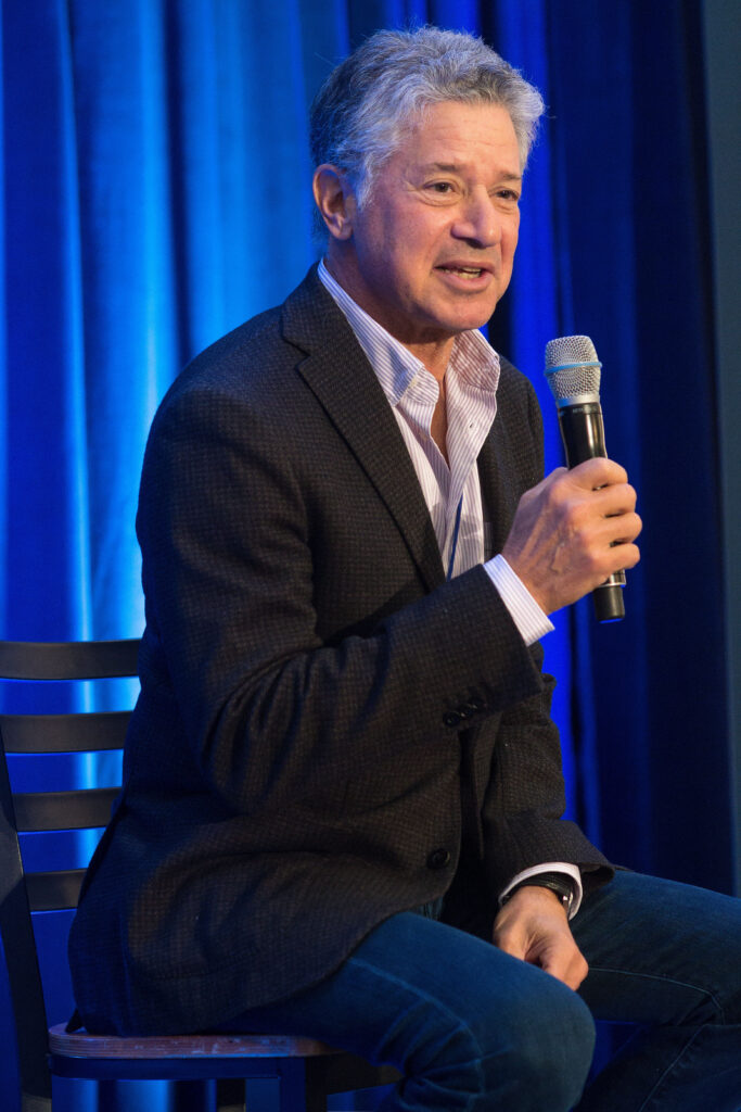 Photograph of a man in his 50s with light skin tone and grey short hair. He is wearing a black suit and thin-striped button up shirt. He is holding a microphone and speaking while looking off camera. 