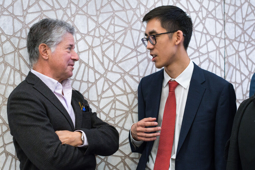 Photograph of two men standing against a white with grey line pattern across it, they are talking to each other. The man on the left is in his 50s and wearing a dark grey suit, the man on the right is in his 20s and wearing a navy suit.