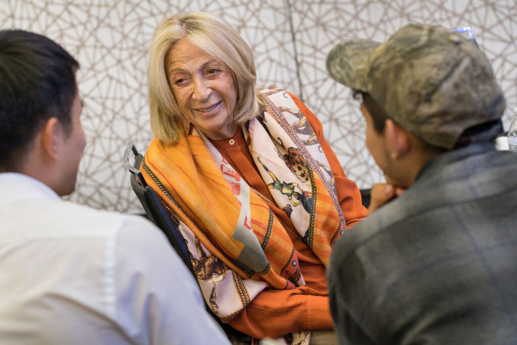 Photograph of three people talking the camera angle is from behind two men who are in their 20s looking directly at the third who is a woman in her 80s. There is a white and grey background behind them.