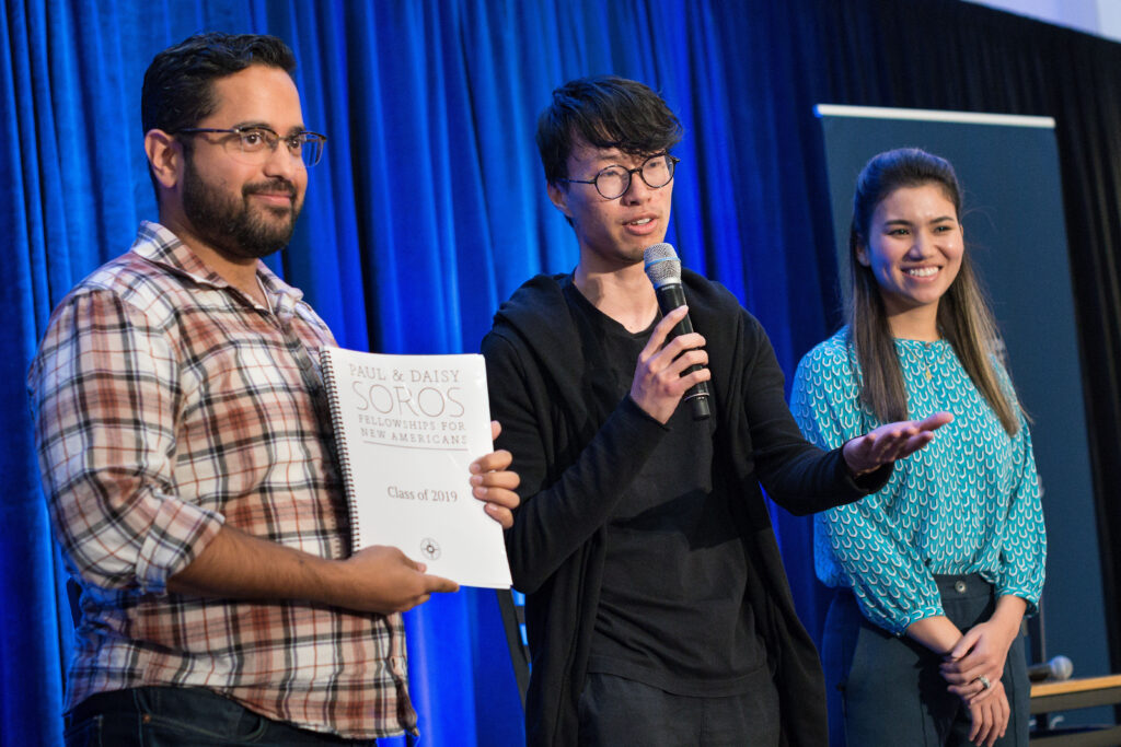 Photograph of three people standing on a stage in front of a blue curtain. The man in the middle holding a microphone is in his 20s. The other two look on, smiling, they are also in their 20s.