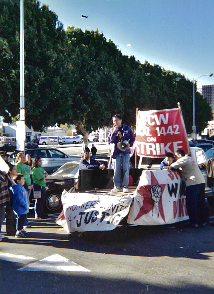 Photograph of a man standing in the bed of a pick-up truck with a bull horn in hand. There are people standing around listening to him speak. The truck has various signs hanging off - not all words are visible "Local 1442 on Strike" can be seen behind the man.