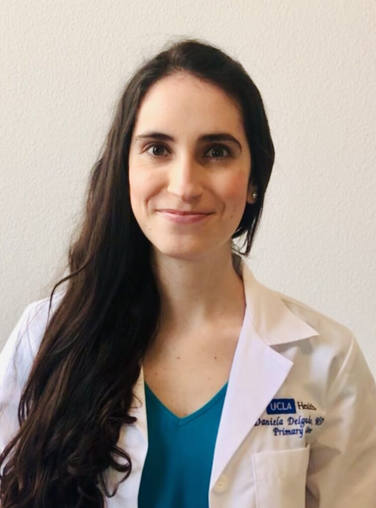 Headshot of a woman in her 20s who has heritage from Colombia, with light skin tone and long black straight hair parted on the side and pulled over her right shoulder. She is wearing a white doctor's coat and "UCLA Health" can be seen embroidered on the left breast, she is also wearing a teal v-neck blouse and pearl stud earrings. She is smiling at the camera. 