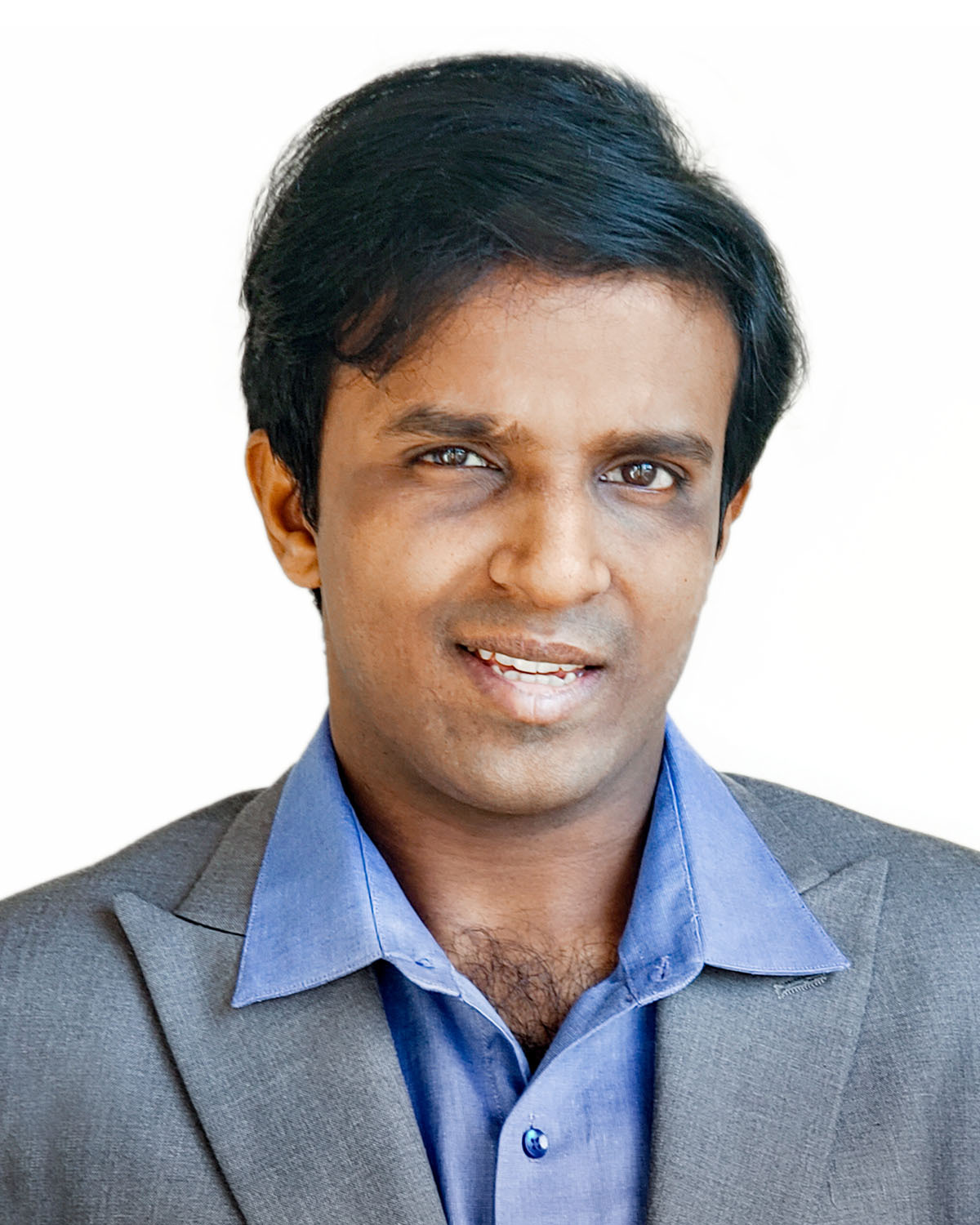 Headshot of a man in his 20s who has heritage from India with medium-dark skin tone and black side combed hair. He is wearing a light grey suit and blue button up shirt. He is smiling at the camera.