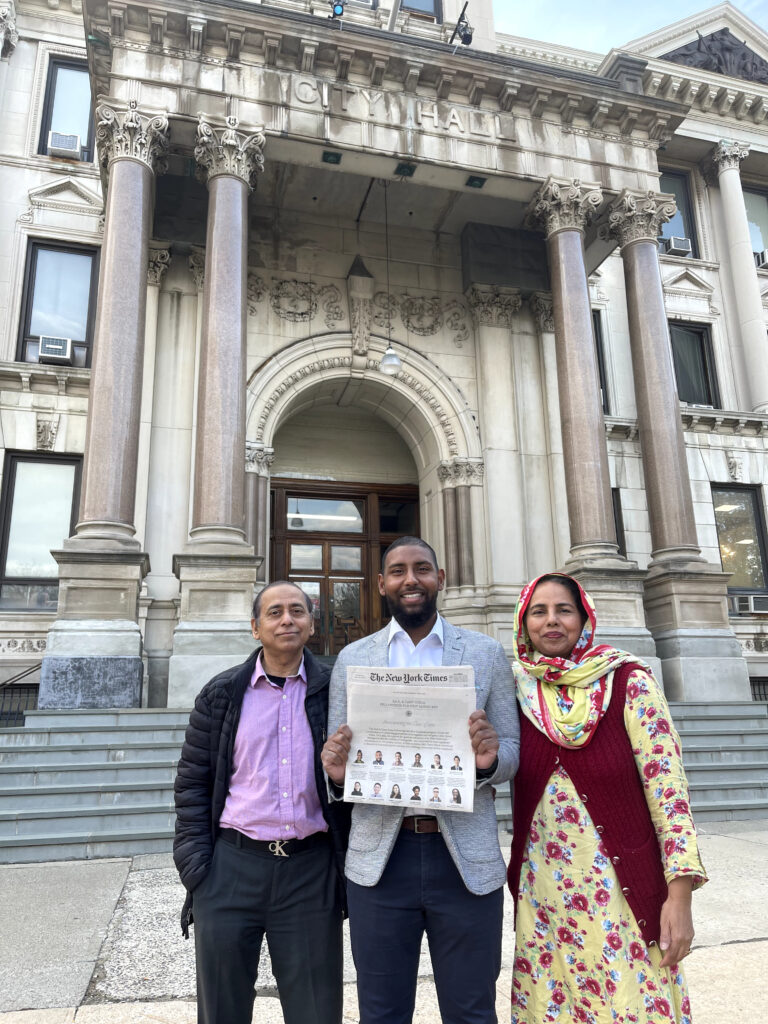 Photograph of three people, the man in the middle is in his 20s he is wearing a grey blazer, white button up shirt and jeans. The people to his left and right are middle-aged. They are standing in front of a stone building with brown marble columns.  