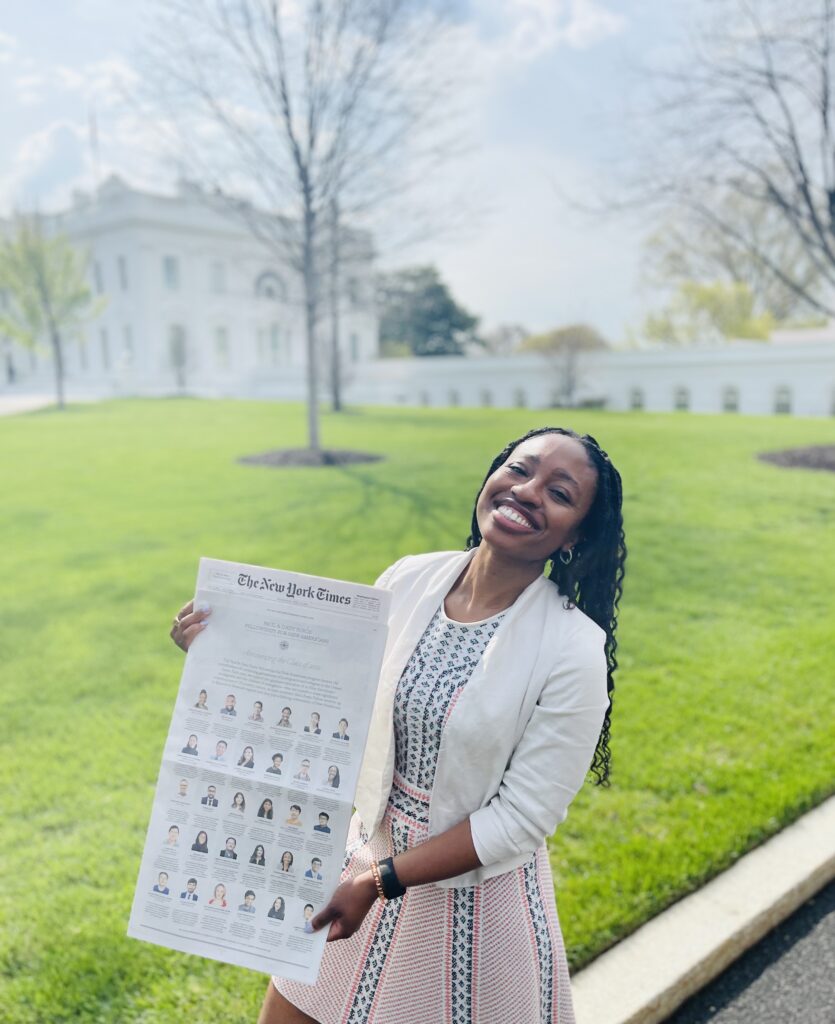 Photograph of a woman in her 20s standing on a sidewalk outside and holding a New York Times, she is wearing a white blazer and a multi-colored patterned dress. Behind her is a green lawn and a large white building in the distance. 