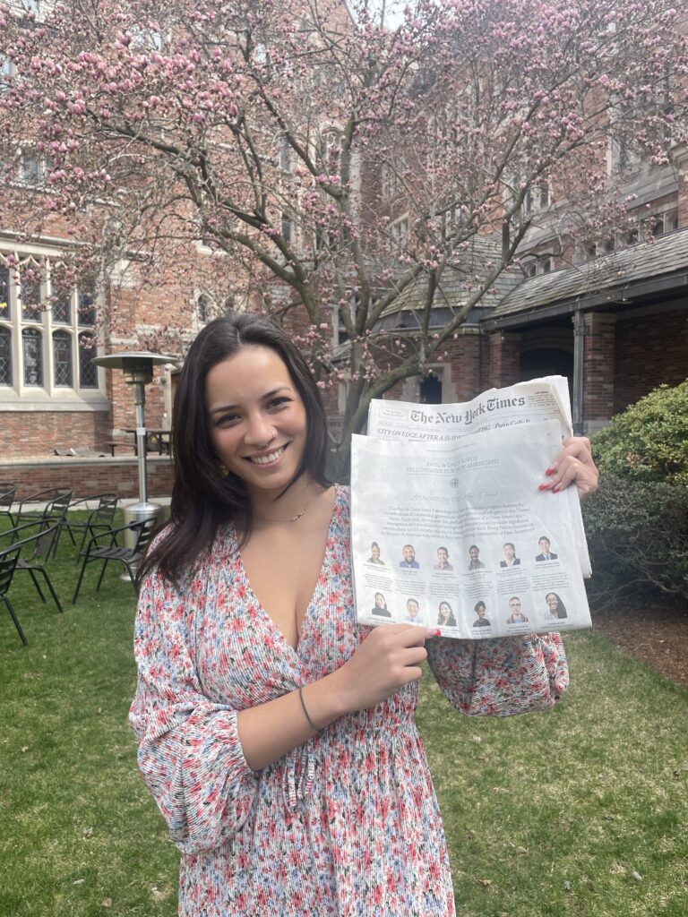 Photograph of a woman in her 20s, standing in the courtyard of a red brick building, cherry blossom tree in bloom. She is holding a New York Times and wearing a floral patterned dress.