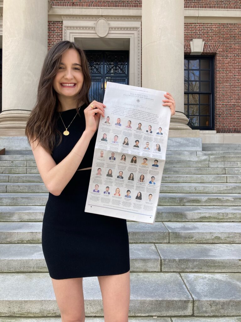 Photograph of a woman in her 20s standing in front of a stone staircase and brick and stone building. She is wearing a black dress and holding a New York Times.