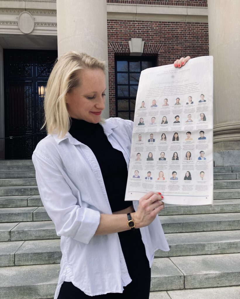 Photograph of a woman in her 20s, standing on a stone staircase outside a stone and brick building. She is holding a New York Times. She is wearing a white button up over a black turtleneck. 