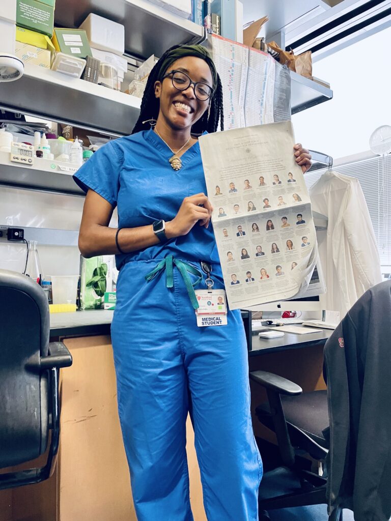 Photograph of a woman in her 20s standing in a lab and holding a New York Times. She is wearing blue scrubs.