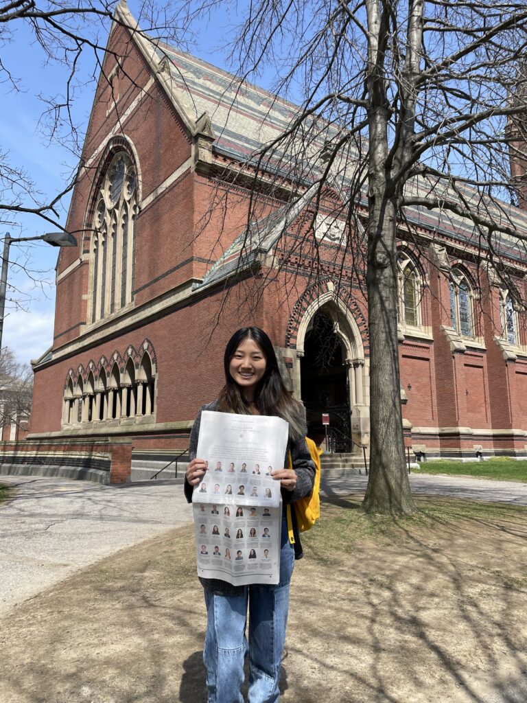 Photograph of a woman in her 20s holding a New York Times and standing in front of a large red brick building. She is wearing a long sleeve top and jeans. 