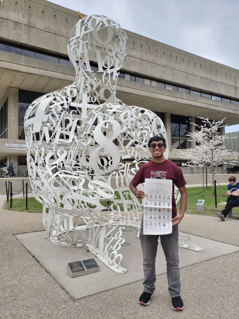 Photograph of a man in his 20s wearing a red MIT t-shirt and jeans and holding a New York Times. He is standing in front of a sculpture in the shape of a human but made of numbers and symbols in white metal. 