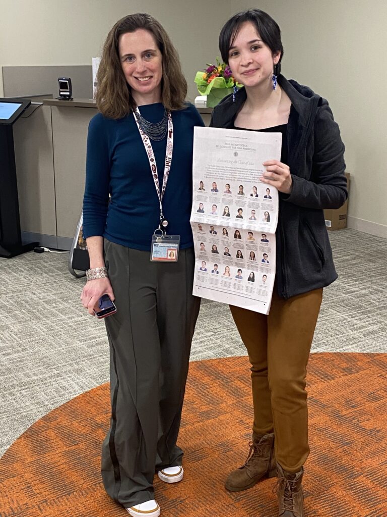 Photograph of two women standing in an office, one woman is in her 20s and is holding a New York Times and wearing a black jacket, black shirt and brown pants. The other woman is in her 40s, wearing a dark teal top, grey pants and a lanyard. 