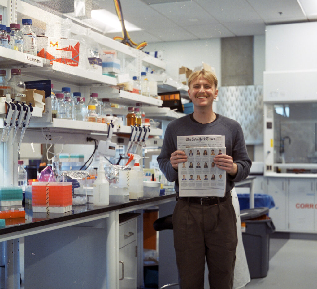 Photograph of a man in his 20s standing in a lab and holding a New York Times. He is wearing a long sleeve grey shirt and black pants.