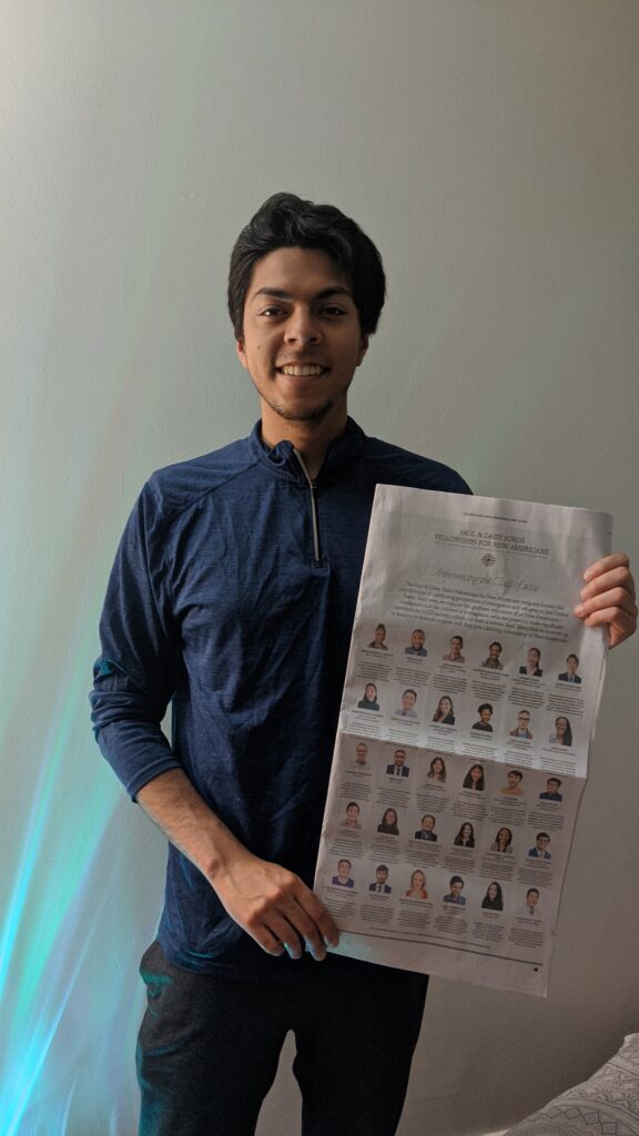 Photograph of a man in his 20s, he is standing against a blank wall background and holding a New York Times. He is wearing a blue half-zip sweater. 