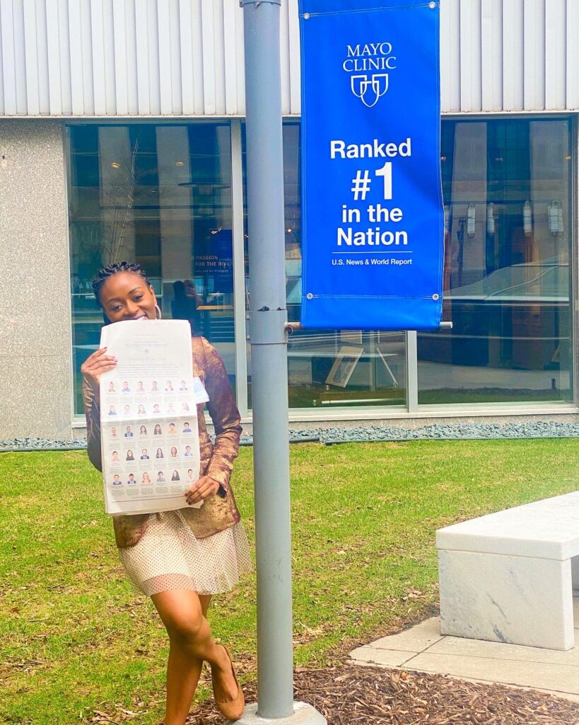 Photograph of a woman in her 20s standing outside next to a lamppost that says "Mayo Clinic" and "Ranked #1 in the Nation". She is holding a New York Times and wearing a mult-colored dress.