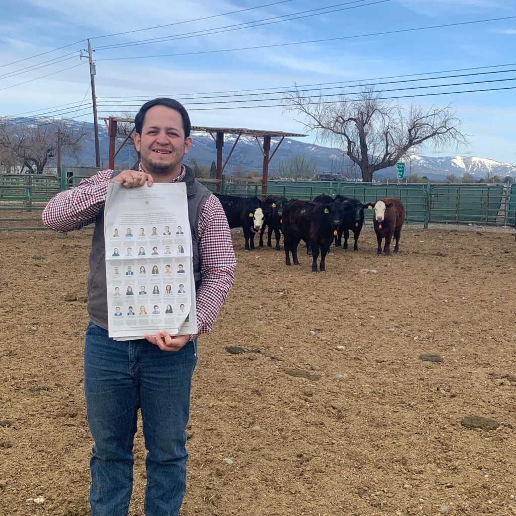 Photograph of a man in his 20s standing on a cattle ranch, many cows can be seen in the background, he is holding a New York Times. He is wearing jeans, a white and red gingham button up and a grey vest. 