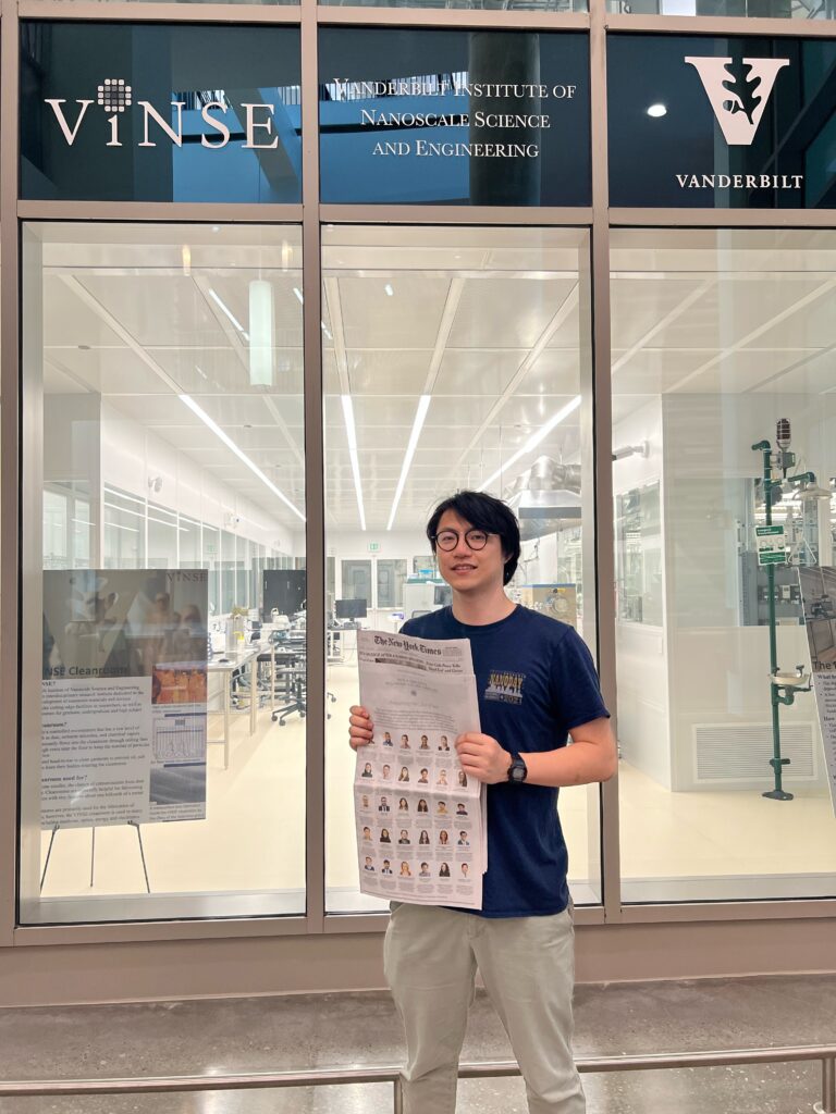 Photograph of a man in his 20s standing in front of a windowed wall that says "Vinse" and "Vanderbilt Institute of Nanoscale Science and Engineering" and "Vanderbilt". He is holding a New York Times, wearing a black t-shirt and light grey pants.