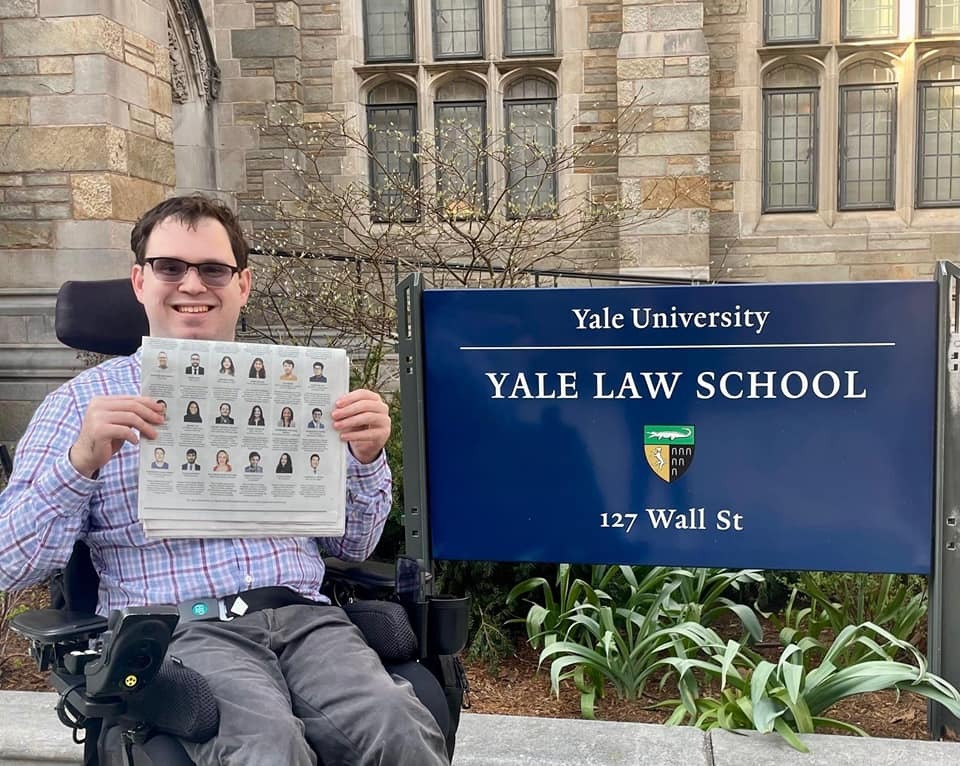 Photograph of a man in his 20s, seated in a power wheelchair and holding a New York Times. He is next to a sign that says "Yale University Yale Law School". He is wearing a red, white and blue tartan button up shirt. 