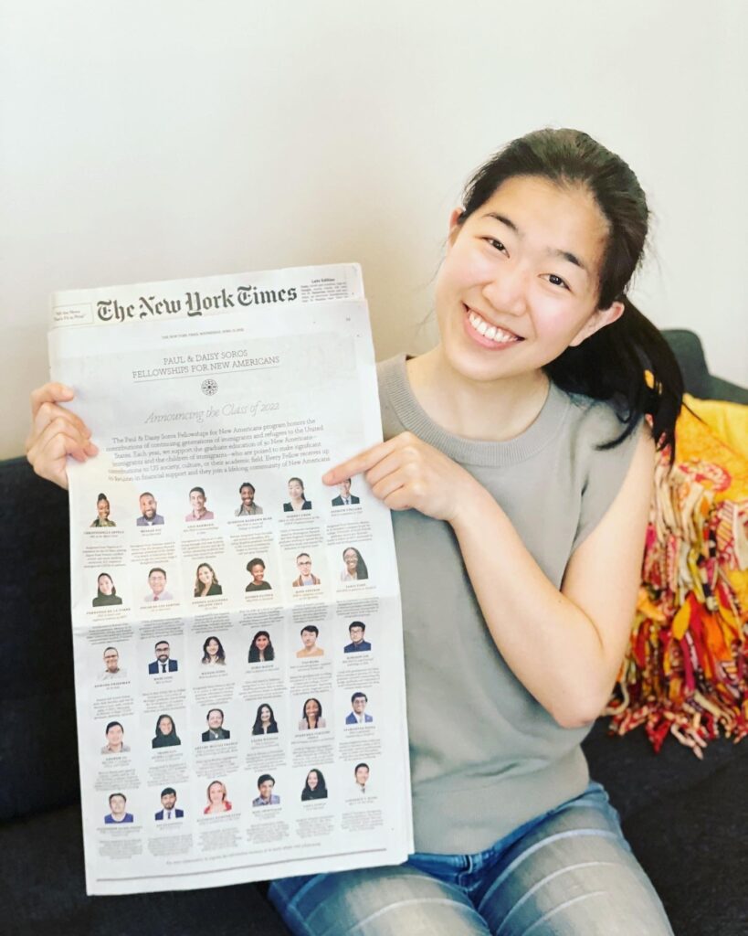 Photograph of a woman in her 20s, sitting on a blue couch and holding the New York Times, she is wearing a grey sleeveless top and jeans.