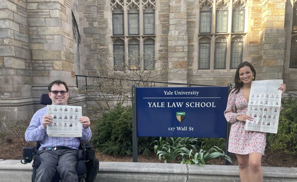 Photograph of a man in his 20s in a power wheelchair wearing a red, white and blue tartan button up, with grey pants and he is holding a New York Times. A woman in her 20s wearing a floral dress and holding a New York Times is also in the photo. They flank a blue sign that reads "Yale University Yale Law School".