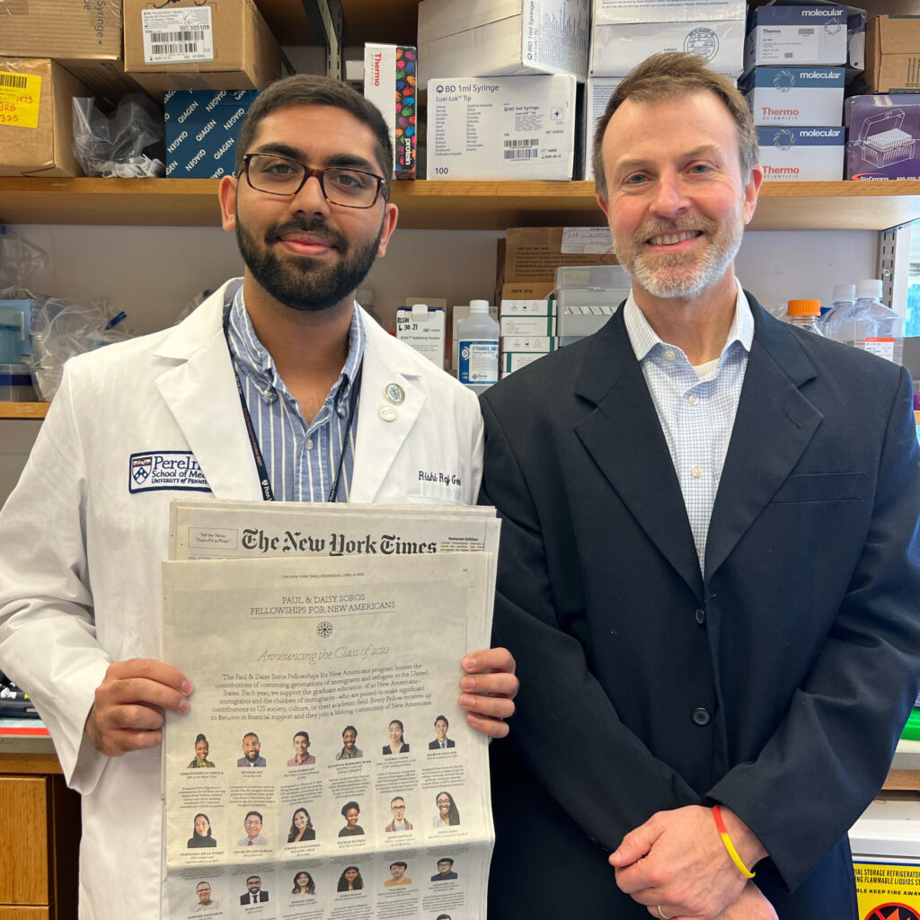 Photograph of two men standing in a lab. The one on the left has a white doctor's coat, blue and white striped button up and is holding the New York Times. The man on the right is in his 50s and has a navy suit with a white and blue gingham button up.